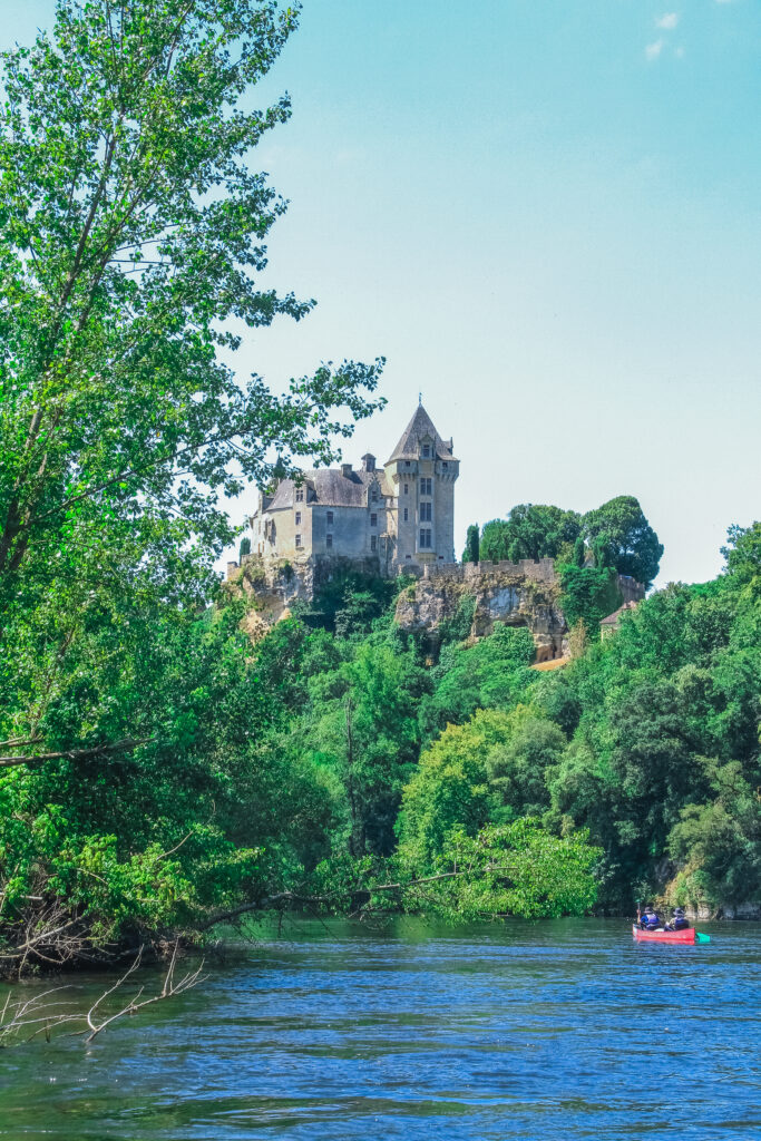 Château en bord de Dordogne entouré par la végétation et visible depuis l’eau par des personnes en canoë rouge.